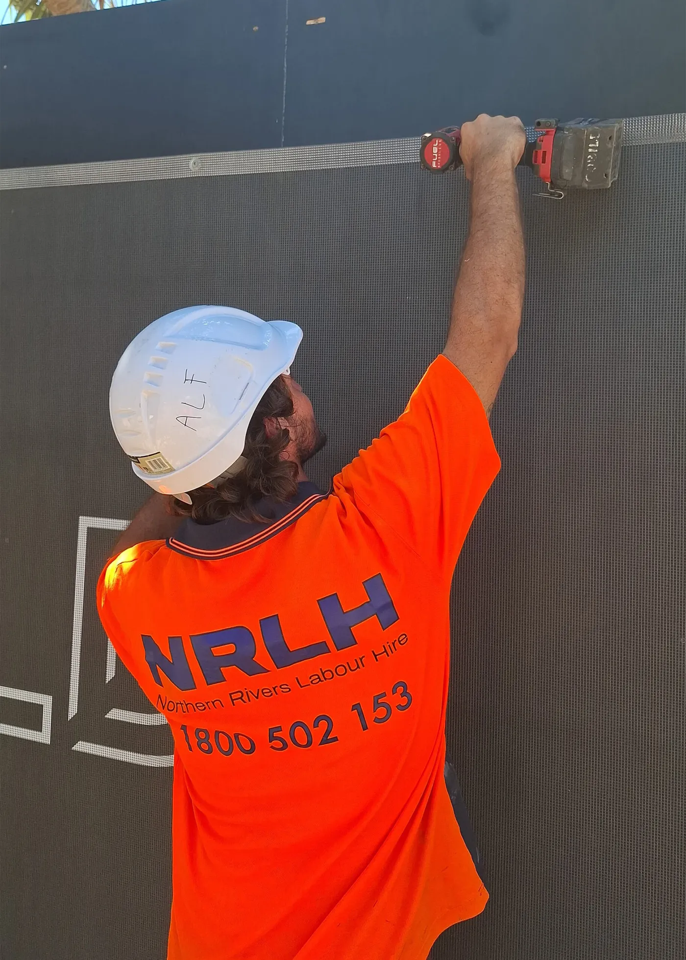 An NRLH labourer in a white hard hat fixing a rail to a dark wall, seen from behind