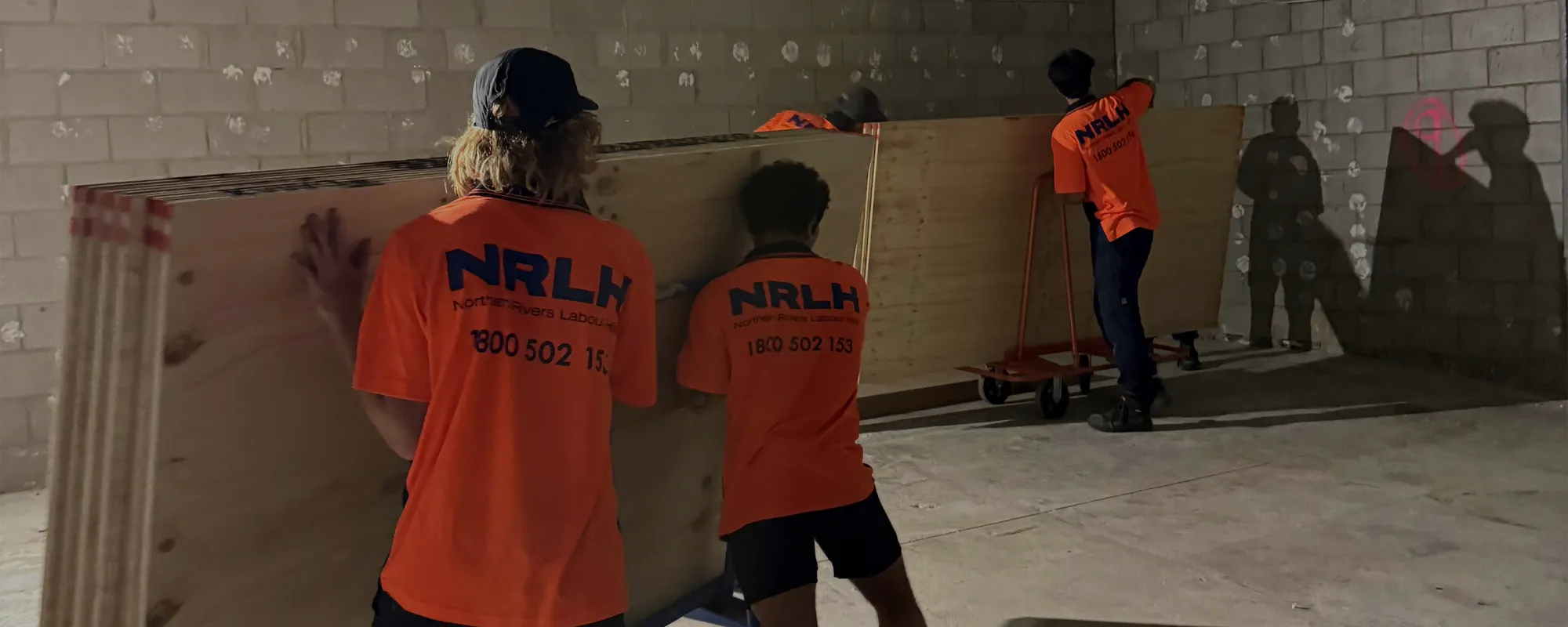 Three NRLH labourers moving sheets of plywood inside a blockwork room