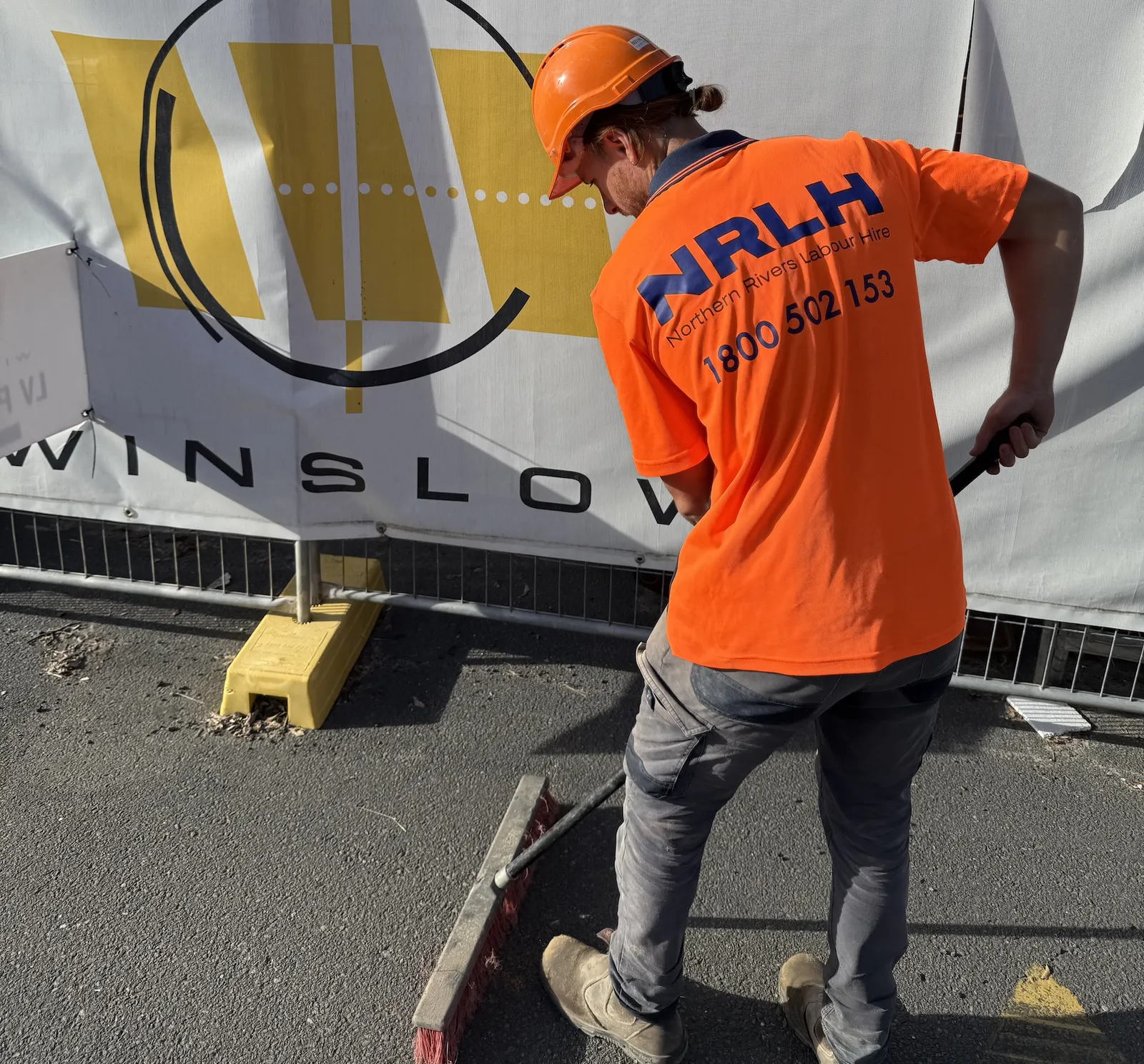 An NRLH labourer in an orange hard hat sweeping beside site hoarding